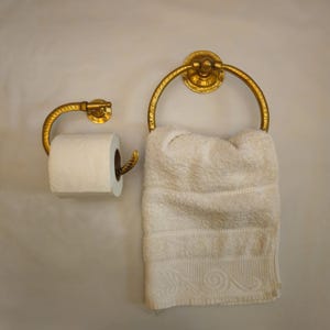Könnte beinhalten: Goldfarbenes Badezimmer-Hardware-Set, einschließlich eines Toilettenpapierhalters und eines Handtuchrings. Der Toilettenpapierhalter hat einen gebogenen Arm, und der Handtuchring hält ein weißes Handtuch. Das Set ist an einer hellen Wand montiert.