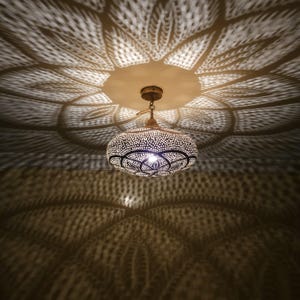 Pode incluir: Luminária pendente ornamentada, com acabamento em latão, projetando sombras florais. A lâmpada tem uma tela redonda e perfurada com um design detalhado. A lâmpada está suspensa no teto.