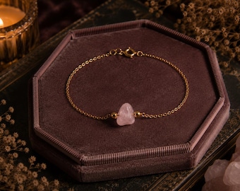 May include: Gold-coloured bracelet with a pale pink rose quartz gemstone. The bracelet is set on a dark burgundy velvet surface. A lit candle and dried flowers are in the background. A beautiful piece of jewellery.