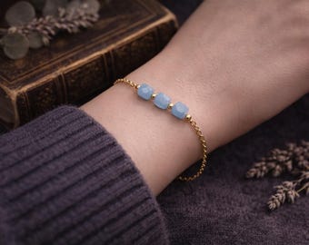 May include: A gold bracelet with three light blue square gemstones. The bracelet is on a wrist, with a dark purple sweater sleeve in the foreground. A vintage book and dried flowers are in the background.