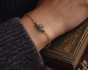 May include: A fine gold chain bracelet with a pale blue-green gemstone. The bracelet has small gold beads and is worn on a wrist. The background includes a dark grey jumper, a vintage book, and dried flowers.