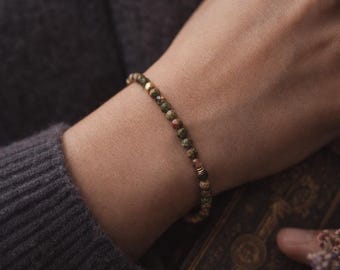 May include: A bracelet made of small, multi-coloured beads, predominantly green, brown, and gold. The bracelet is worn on a wrist, set against a dark grey sweater. A subtle piece of jewellery.