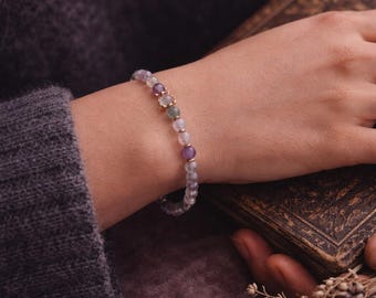 May include: A delicate beaded bracelet featuring purple, pink, and gold elements. The bracelet is displayed on a wrist, with a book and dried flowers in the background. The bracelet incorporates small, round beads in shades of purple and pink, complemented by gold-toned beads.