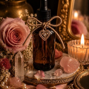 May include: An amber glass dropper bottle with a black cap, adorned with a jute twine bow and a gold-toned heart-shaped charm featuring a central clear stone. The bottle is cylindrical with a smooth surface and a standard glass pipette dropper.