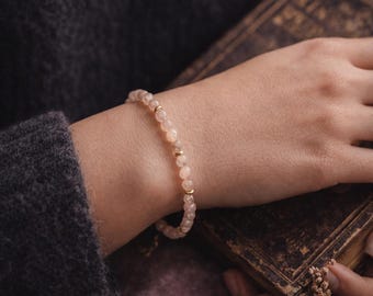 May include: A delicate bracelet with small, pale pink beads and gold-coloured spacers. The bracelet is worn on a wrist, with a dark grey jumper in the background. The bracelet is a piece of jewellery.