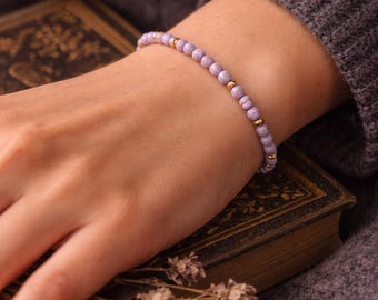 May include: A delicate bracelet featuring small, light purple beads interspersed with gold-coloured accents. The bracelet is displayed on a wrist, with a vintage book and dried flowers in the background. A simple and elegant piece of jewellery.