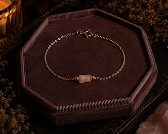 May include: A fine gold chain bracelet featuring a raw rose quartz gemstone. The bracelet is presented on a dark burgundy velvet jewellery box. A lit candle and dried flowers are in the background.