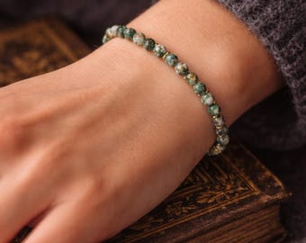 May include: A bracelet featuring green and white speckled beads with gold-coloured spacers. The bracelet is worn on a wrist, resting on a vintage book. The background is a grey sweater.