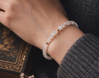 May include: A delicate bracelet featuring small, faceted, clear beads strung together. The bracelet is accented with small gold-coloured beads. The jewellery is worn on a wrist, with a dark grey sweater in the background.