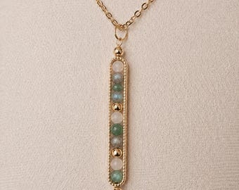 May include: A gold-coloured necklace with a vertical pendant. The pendant features a series of colourful, round gemstones in shades of white, green, and blue. The necklace chain is delicate and also gold-coloured.