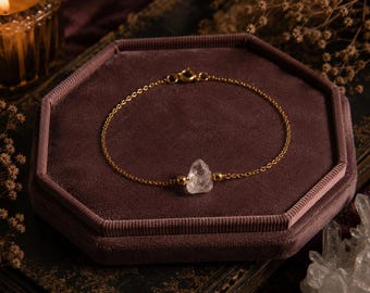 May include: A gold chain bracelet featuring a clear quartz crystal pendant. The bracelet is presented on a hexagonal velvet jewellery box. The background includes a lit candle and dried flowers.