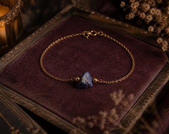 May include: A gold chain bracelet with a triangular lapis lazuli stone. The bracelet is presented on a dark purple velvet surface. The gold chain and stone are the focal point, with a warm light in the background. A classic jewelry piece.