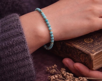May include: A turquoise beaded bracelet with gold detailing. The bracelet is on a wrist, with a vintage book and dried flowers in the background. The bracelet features small, round turquoise beads and gold-coloured spacers.
