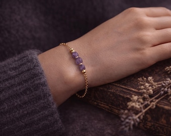 May include: A gold bracelet with three purple amethyst beads. The bracelet is on a chain. The background includes a dark grey sweater and a vintage book. The bracelet is resting on a person's wrist.