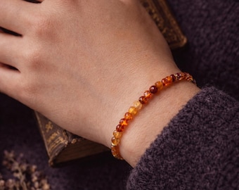 May include: A bracelet made of small, round amber-coloured gemstones. The bracelet is worn on a wrist, with a dark purple sweater sleeve visible. The gemstones vary in colour from light yellow to deep orange and red.