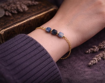 May include: A gold bracelet with three blue gemstone beads. The bracelet is worn on a wrist, with a dark purple sweater in the background. Dried lavender sprigs and a vintage book are also visible.