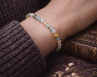 May include: A bracelet featuring a mix of pastel-coloured beads, including yellow, blue, and white, with small gold-coloured beads. The bracelet is worn on a wrist, with a dark purple sweater in the background. The bracelet is approximately 18 cm in circumference.