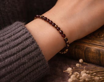 May include: A bracelet with small, round, dark red gemstones and gold-coloured beads. The bracelet is worn on a wrist, with a dark grey sweater in the background. The bracelet is a piece of jewellery.