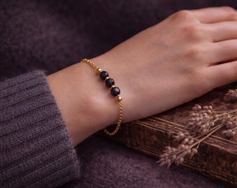 May include: A gold chain bracelet featuring three black gemstone beads. The bracelet is displayed on a wrist, with a dark purple sweater in the background. The bracelet is approximately 18 cm in length.