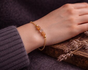 May include: A gold bracelet featuring three orange gemstones. The bracelet is worn on a wrist, resting on a book. The background is a dark grey sweater.