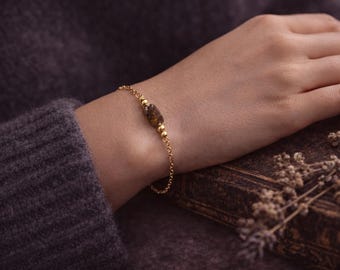 May include: A gold bracelet featuring a chain and a central, oblong, brown and gold gemstone. The bracelet is on a wrist, with a dark grey sweater sleeve in the background. The bracelet is a delicate piece of jewellery.