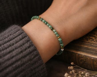 May include: A delicate bracelet with small, round, green beads and gold-coloured accents. The bracelet is worn on a wrist, with a dark grey sweater sleeve visible. The background includes a book and dried flowers.