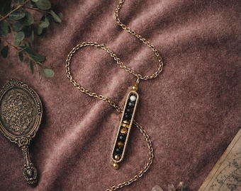 May include: A gold necklace featuring a rectangular pendant adorned with black, gold, and white beads. The chain lies on a mauve fabric. A vintage hand mirror and a sprig of greenery are visible in the background.