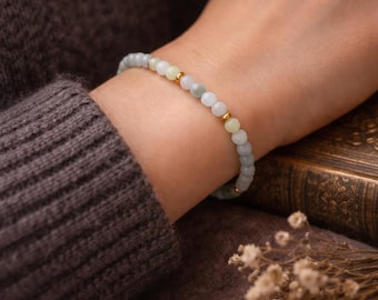May include: A delicate beaded bracelet with small, round, pale green and white stones and gold-coloured beads. The bracelet is worn on a wrist, with a dark brown sweater sleeve in the background. The bracelet is a piece of jewellery.