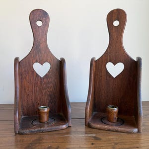 Puede incluir: Dos candeleros de madera marrón oscuro. Cada uno tiene un asa con un agujero, un recorte en forma de corazón y una pequeña copa de metal para velas. Los candeleros se exhiben sobre una superficie de madera.