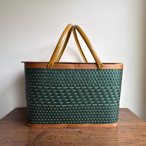 May include: A rectangular picnic basket with a woven dark green pattern and wooden trim. The basket has two arched, light brown handles. The basket is sitting on a wooden table.
