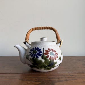 Pode incluir: Uma chaleira de cerâmica com acabamento branco salpicado, com desenhos florais azuis e vermelhos e uma alça tecida. A chaleira tem um bico e uma tampa e fica em uma superfície de madeira.