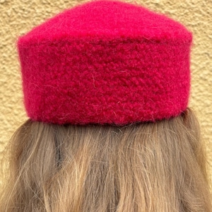 May include: A vibrant, bright red pillbox hat with a fuzzy, textured surface. The hat features a flat top and straight, structured sides, showcasing a dense, fibrous material that creates a soft, tactile appearance throughout the entire piece.