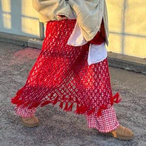 Könnte beinhalten: Eine Person trägt eine beige Jacke, ein weißes Hemd, einen roten Häkelrock mit Fransen und eine rot-weiß karierte Hose. Die Person trägt auch braune Wildlederstiefel.