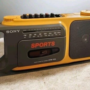 May include: A retro yellow and grey Sony Sports radio cassette player. The radio features a handle with an orange accent, a speaker, and the word "SPORTS" in red. It has a tuner dial and function buttons.