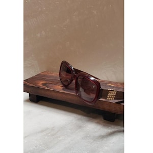 May include: Burgundy-colored sunglasses with a square frame rest on a dark wooden display. The sunglasses have a gold-toned accent on the temples. The display is a small, rectangular wooden shelf.