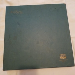 Pode incluir: Uma caixa de jogo de tabuleiro quadrada, de cor verde-azulado escuro com acabamento fosco. O canto superior esquerdo apresenta a palavra Scrabble em letras douradas. O canto inferior direito exibe um logotipo quadrado dourado com S&R e GAMES.