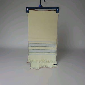 May include: A cream-colored scarf with blue and black horizontal stripes near the bottom. The scarf has a fringed edge and is hanging on a black hanger. The scarf is made of a soft material.