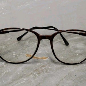 May include: Round-framed eyeglasses with a tortoiseshell pattern on the rims and temples. The lenses are clear, and the bridge and arms are dark brown. The glasses are resting on a gray surface.