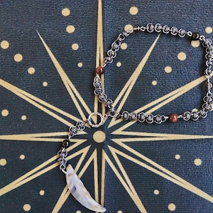 May include: A handmade necklace featuring a silver chain with a pendant shaped like a tooth. The necklace incorporates small, round, brown and black beads. The jewelry is displayed on a dark blue surface with a gold starburst design.