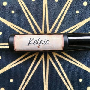 May include: A black roller bottle with the word "Kelpie" in script on a label. The label has a vintage design with a brown and cream color scheme. The bottle is set against a dark blue background with a gold starburst pattern.