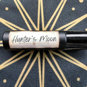 May include: A black roll-on bottle with the words "Hunter's Moon" on a label. The label has a decorative border and a small floral design. The bottle is set against a dark background with a gold starburst pattern.