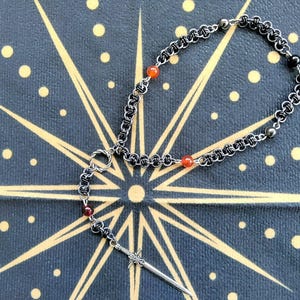 May include: A silver-toned chainmail bracelet with red and black beads, and a sword pendant. The bracelet is displayed on a dark blue surface with a gold starburst design and gold dots.