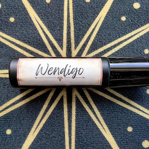 May include: A black essential oil roller bottle with a label that reads "Wendigo" in a script font. The label has a light brown background with a decorative border. The bottle is set against a dark blue and gold patterned surface.