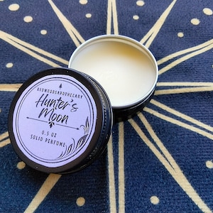 May include: A black and silver solid perfume tin with the text "Hunter's Moon" and "0.5 oz Solid Perfume" on a light purple label. The tin is open, revealing a white solid perfume. The background is a dark blue fabric with a white starburst pattern.