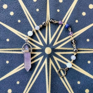 May include: A handcrafted crystal pendant necklace featuring a faceted, lavender-colored crystal point. The necklace includes a chain of small, colorful beads in shades of purple, blue, and black. The pendant hangs on a gold-colored chain. The background is a dark blue surface with a gold starburst design.