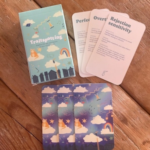 May include: A card game called "Traitspotting" with a light blue box showing a cat on a cloud, a rainbow, and the game's title. Several cards are visible, with titles such as "Rejection Sensitivity" and "Perfectionism".