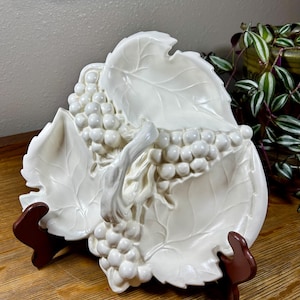 May include: A white ceramic serving dish with a grape and leaf design. The dish is supported by a brown stand. The grapes and leaves are raised, creating a textured surface. The dish is likely used for serving fruit or snacks.