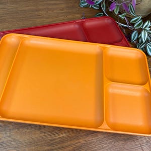 May include: Two plastic divided trays. The top tray is orange, and the bottom tray is red. The orange tray has four compartments: a long, curved section and three square sections. The red tray is visible beneath the orange tray.