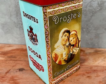 May include: A rectangular metal tin with a red lid, featuring light blue and olive green panels. The tin displays the text Droste's Cocoa and Cacao & Chocolade, along with an illustration of two figures and a red crest logo on the side.