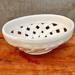 May include: An oval-shaped white ceramic bowl featuring a decorative lattice weave design on its sides. The bowl has a smooth, glossy finish and a solid, continuous rim that encircles the open, woven-style structure of the main body.